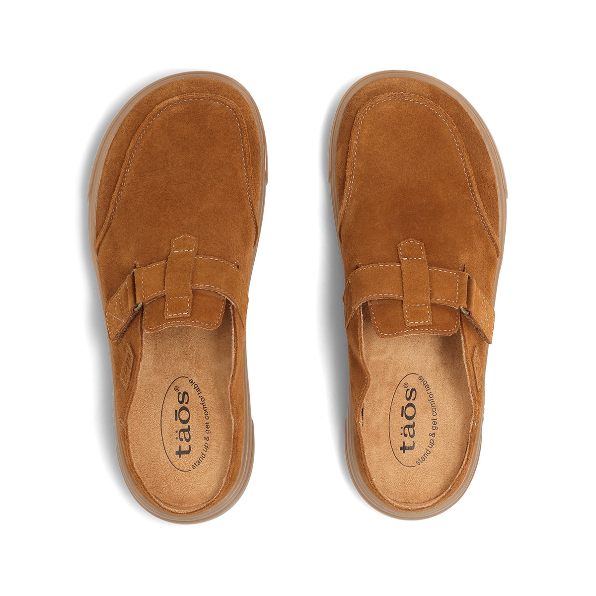 A pair of tan suede clogs shown from a top-down view on a white background. The shoes feature a single strap across the instep with a hook-and-loop closure and a visible 'taos' logo printed on the insole.