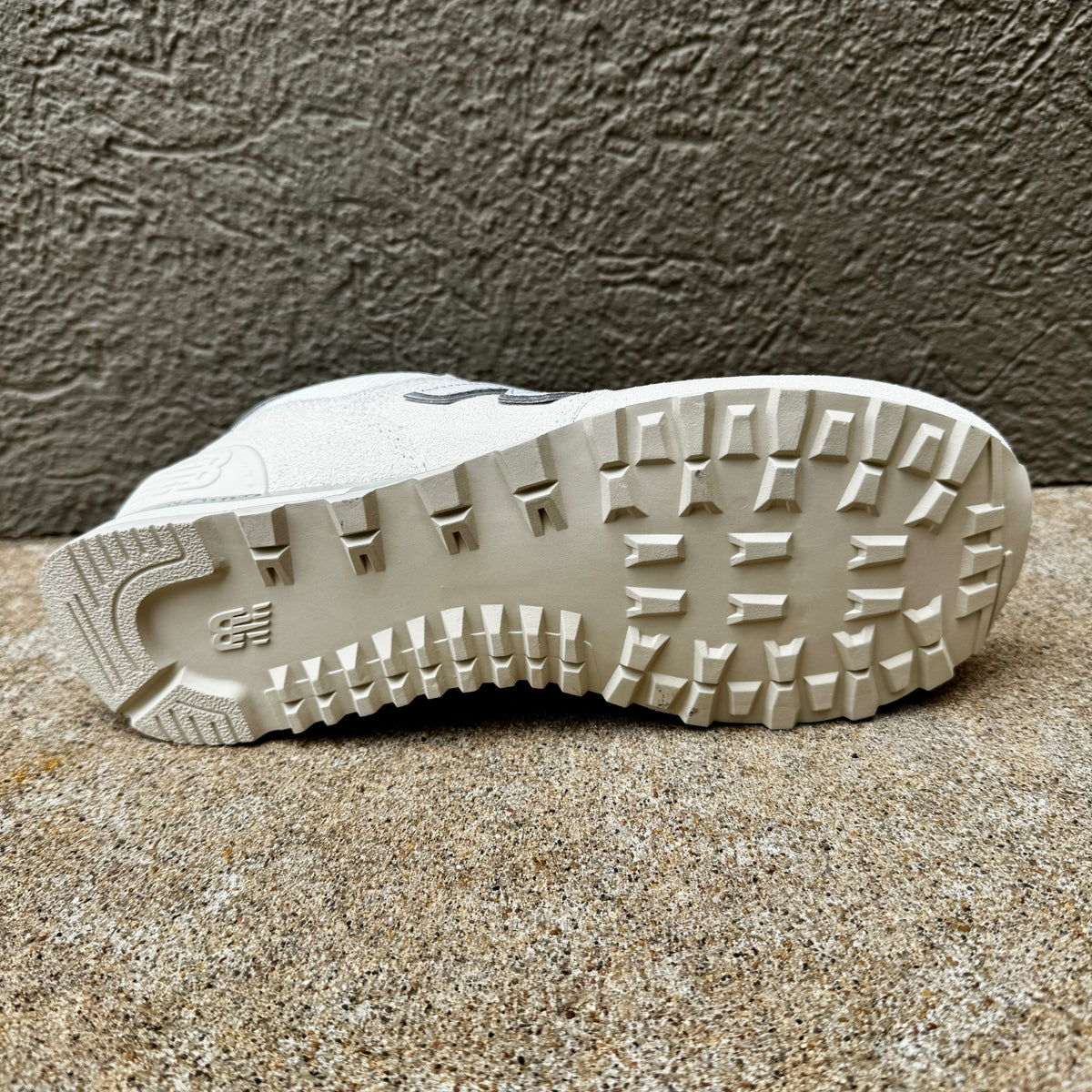 A close-up of the outsole of a white New Balance sneaker, showing the tread pattern and embossed 'NB' logo, resting on a textured concrete surface.