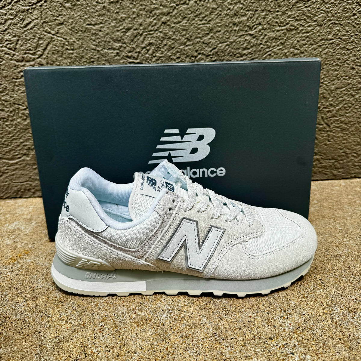 A single New Balance sneaker in white and light gray, positioned in front of its dark gray shoebox on a textured tan surface.