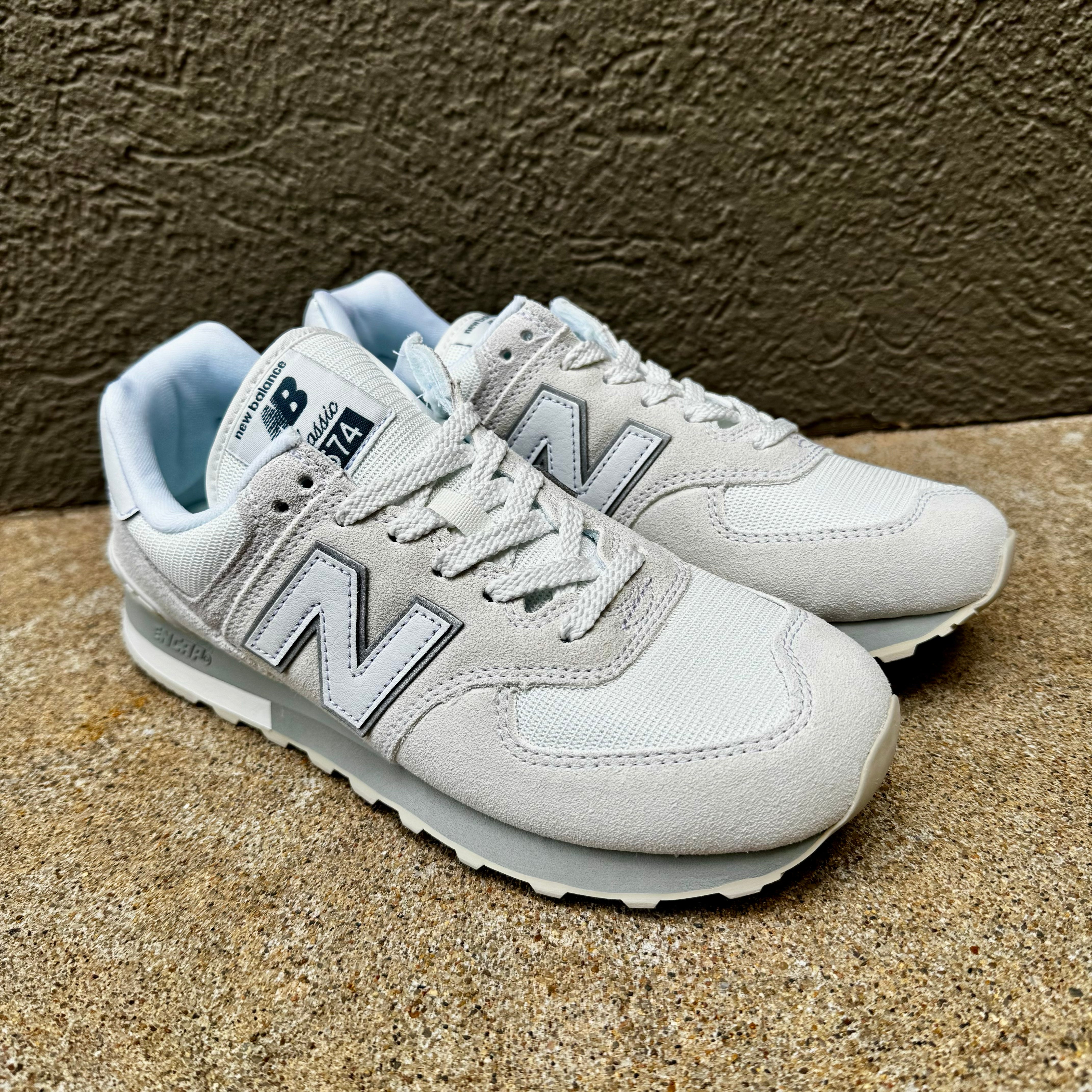 A pair of New Balance 574 sneakers in light gray and white, featuring the signature 'N' logo, lace-up closure, and light blue lining, set against a textured brown background.