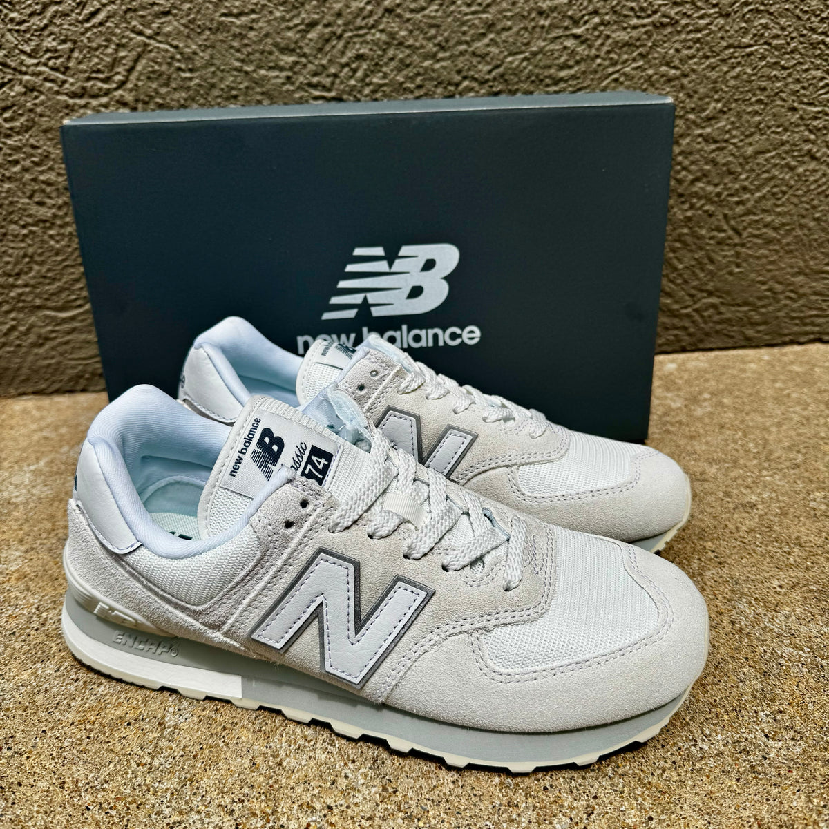 A pair of New Balance 574 sneakers in white and light gray, shown with their dark gray New Balance shoebox on a textured tan surface.