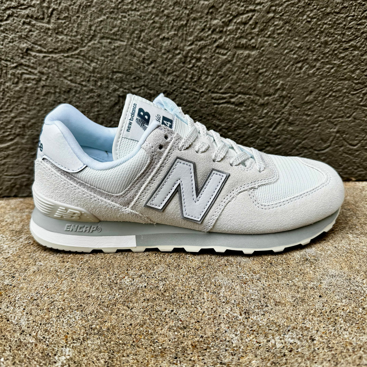A light gray and white New Balance sneaker with a light blue lining, shown in a side profile against a textured brown wall and concrete floor.