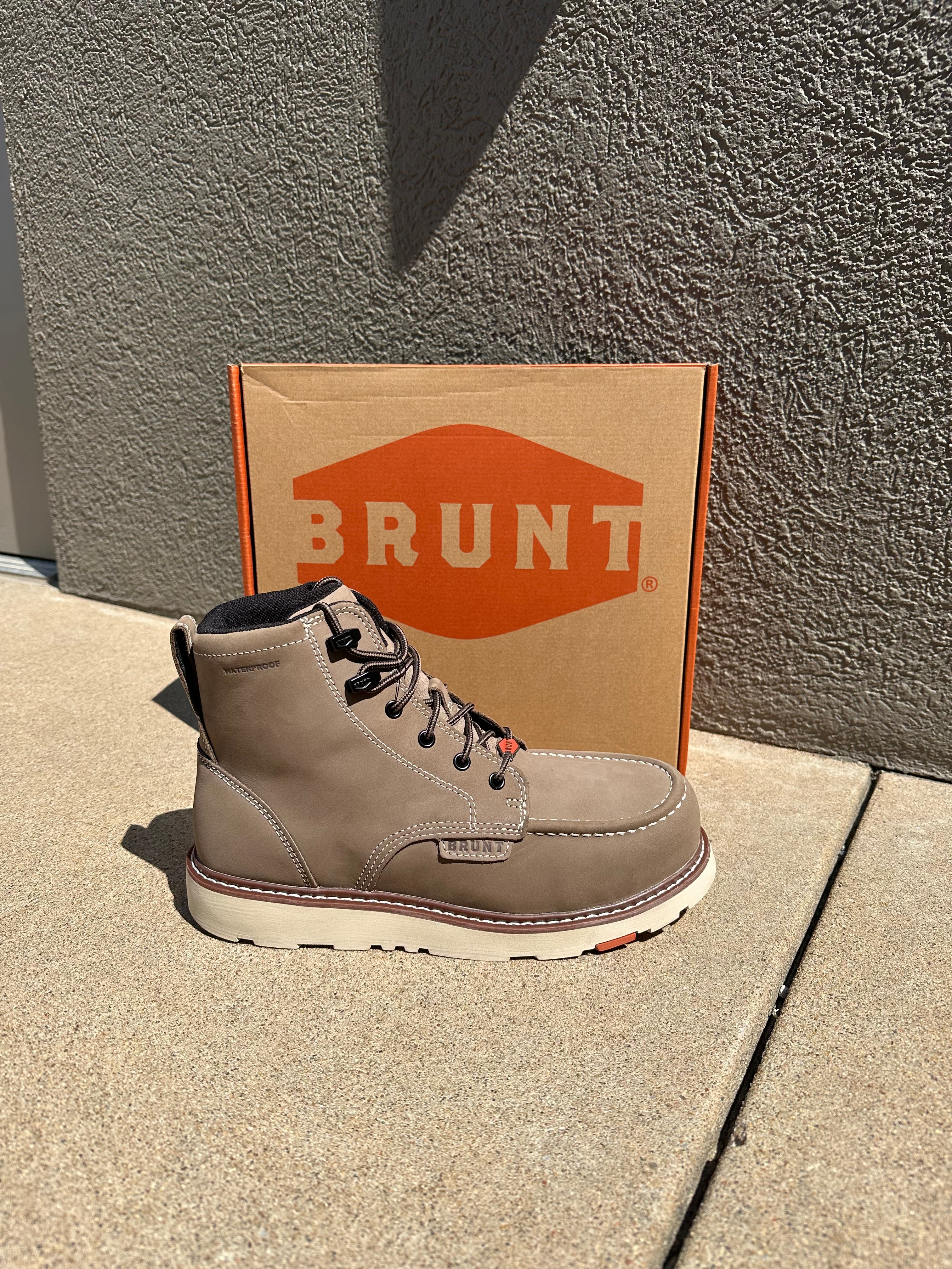 A light gray suede high-top shoe by Brunt, shown next to its brown cardboard box with a prominent orange logo, set against a concrete background.