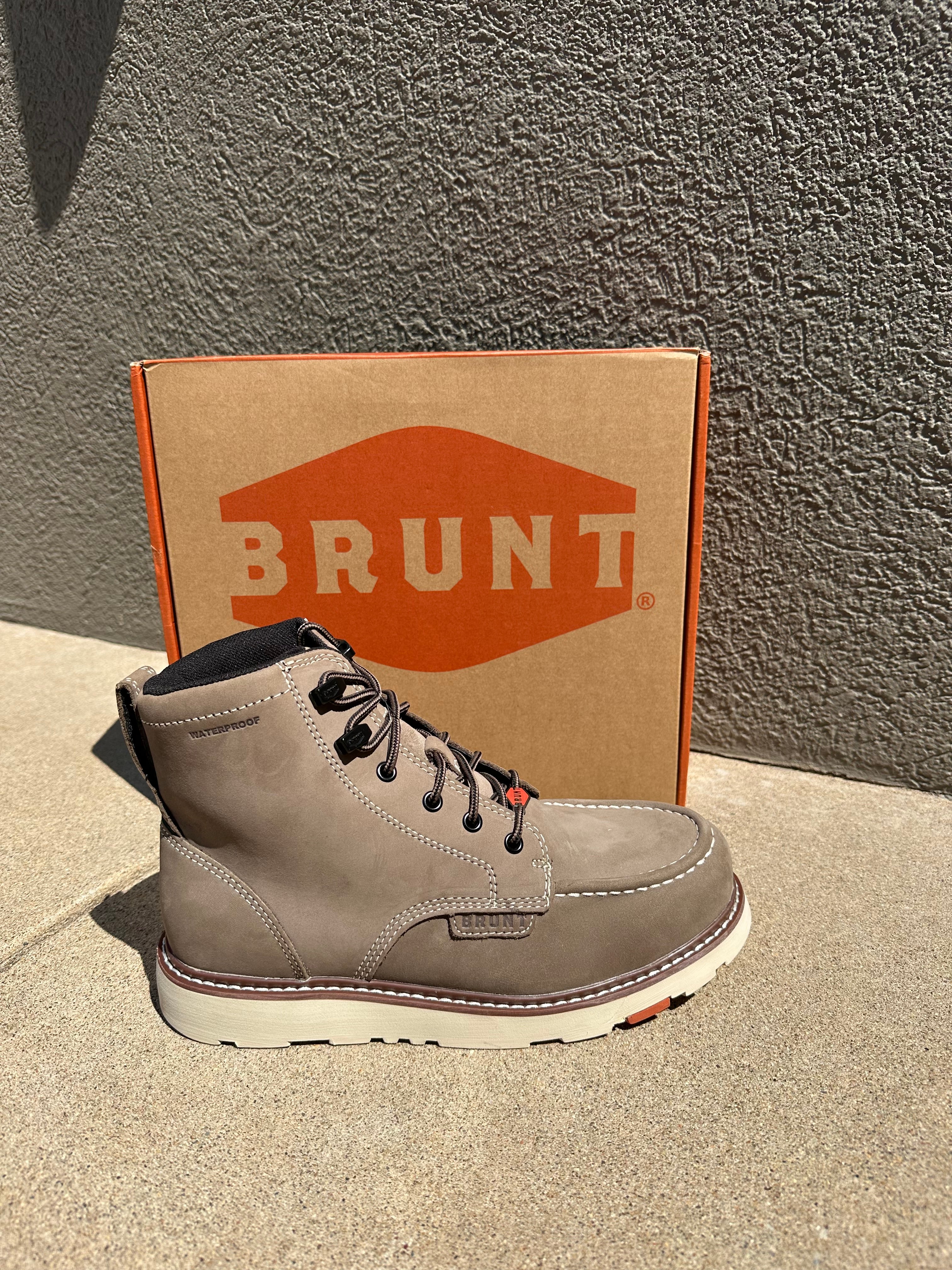 A light gray suede lace-up boot by Brunt, shown next to its brown cardboard box with a prominent orange logo, set against a textured gray concrete background.