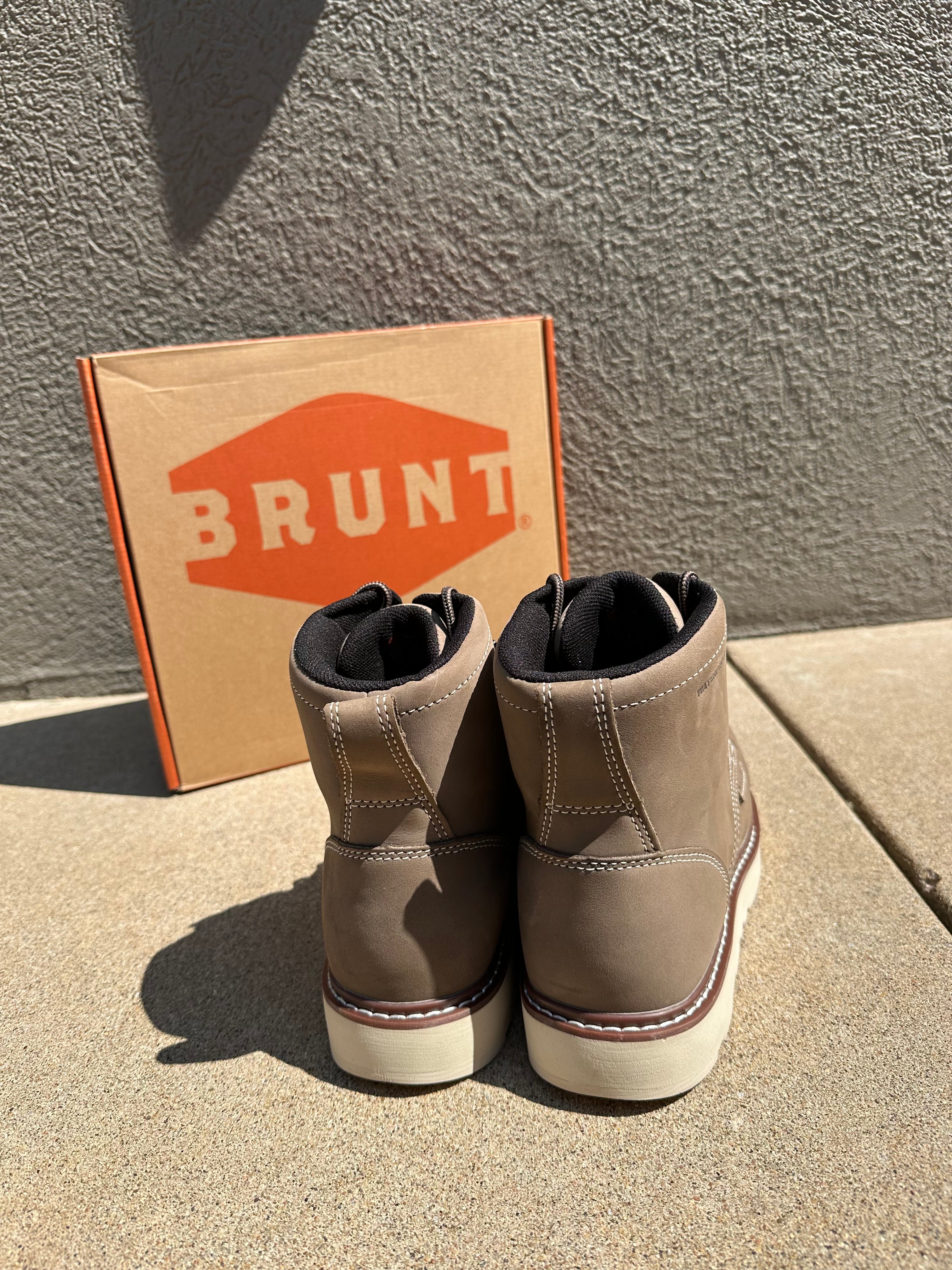 A pair of taupe-brown suede slip-on sneakers with white soles and a thin red stripe, shown next to their cardboard box featuring the orange and white BRUNT logo, set on a concrete surface in bright sunlight.