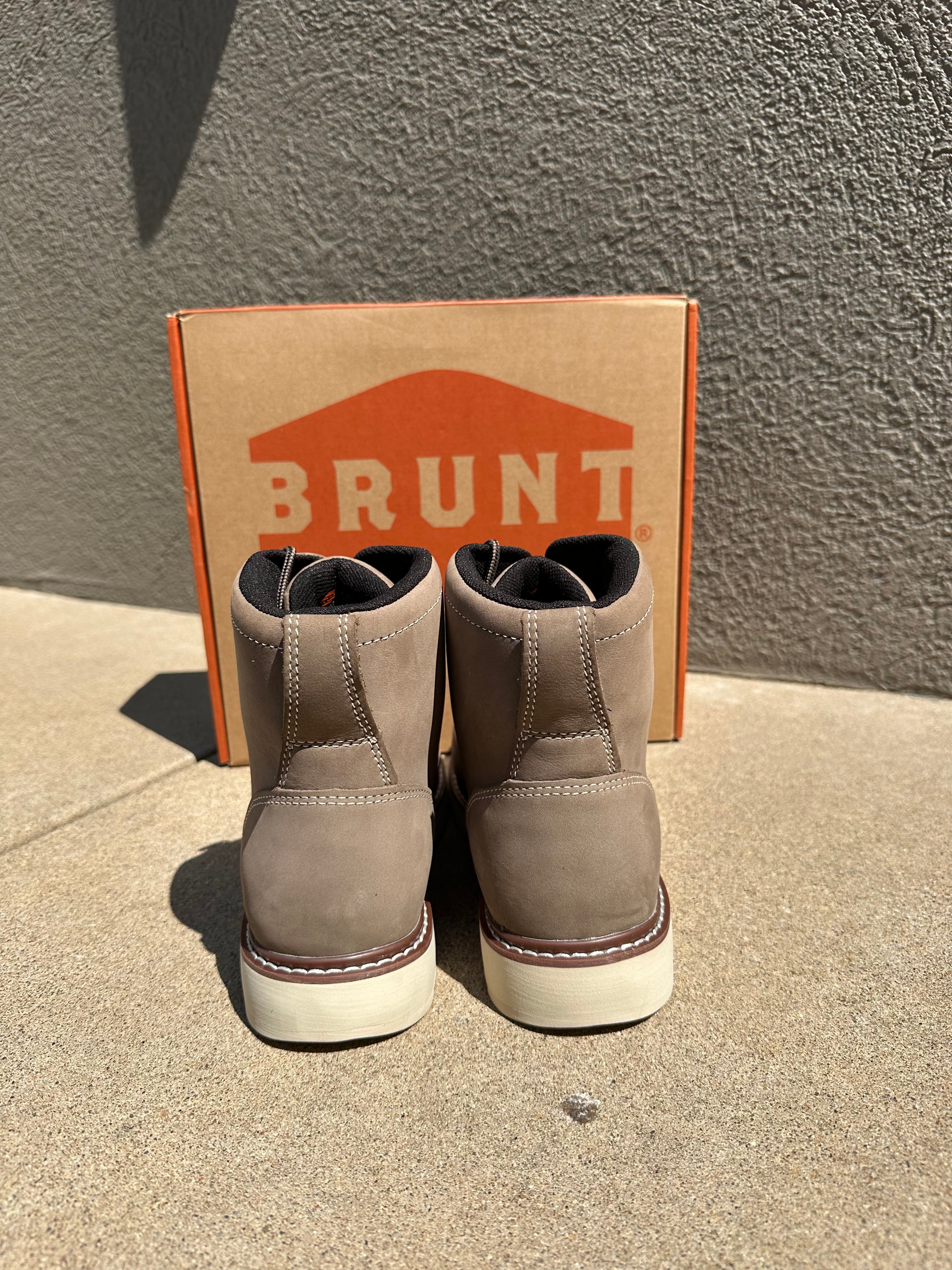 A pair of taupe suede ankle boots with white soles, displayed on top of their brown cardboard Brunt box on a concrete surface.