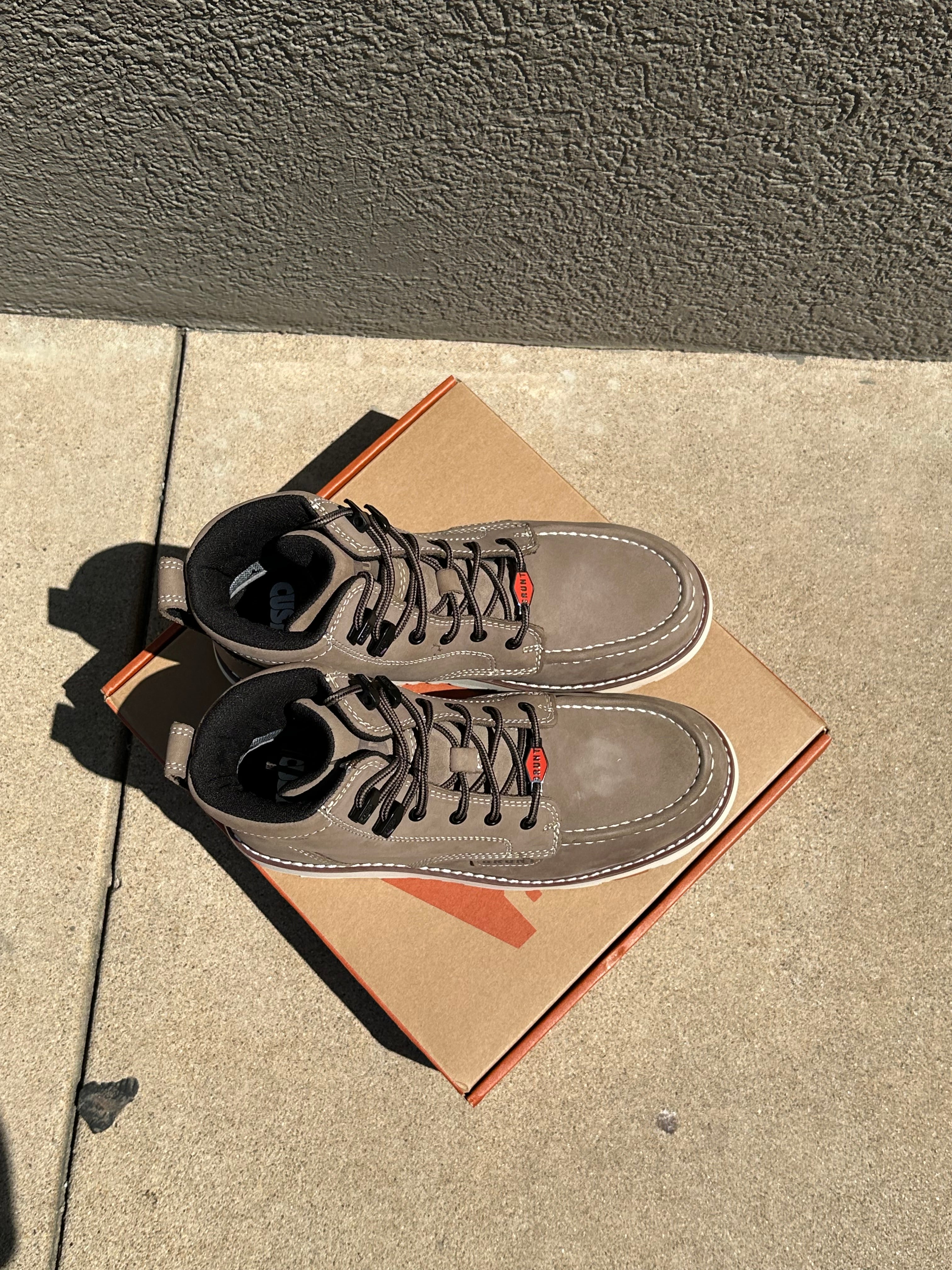 A pair of taupe-brown suede high-top sneakers with black laces, resting on top of their brown cardboard box on a concrete sidewalk.