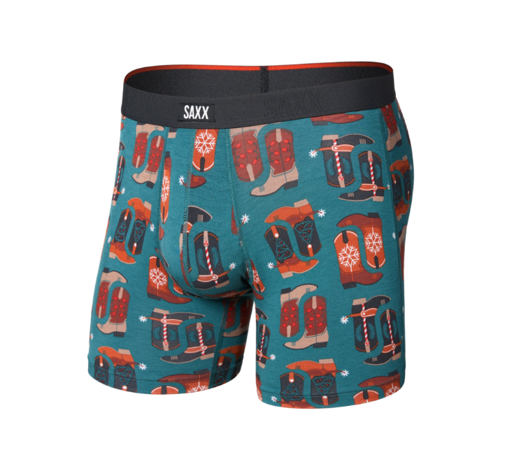 A pair of men's boxer briefs by SAXX, featuring a teal blue background with an all-over print of red and brown cowboy boots, black hats, and small white snowflakes. The underwear has a black waistband with the brand name 'SAXX' in white.